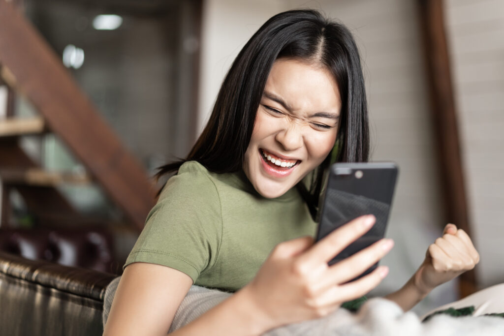 smiling asian woman looking satisfied making fist pump reading mobile phone sitting home